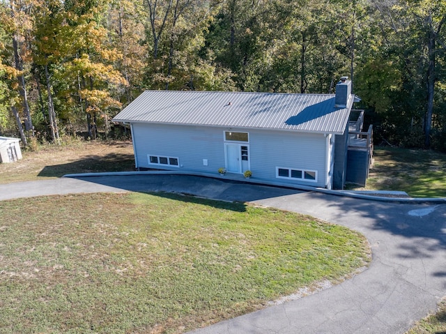 ranch-style house with aphalt driveway, a chimney, metal roof, a view of trees, and a front lawn