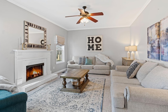 carpeted living room with a warm lit fireplace, ceiling fan, baseboards, and crown molding