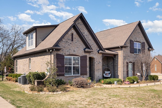 french country inspired facade featuring a front yard, central AC unit, brick siding, and a shingled roof