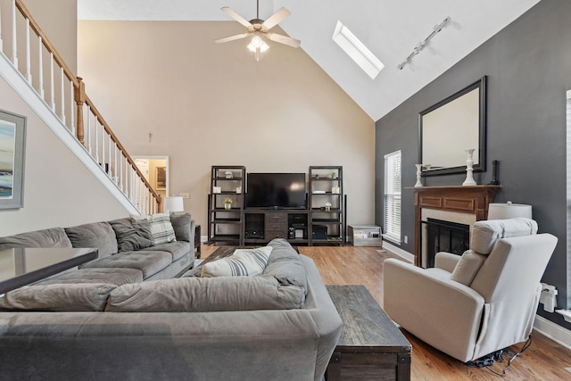 living area with a skylight, stairs, light wood-style floors, a fireplace, and high vaulted ceiling