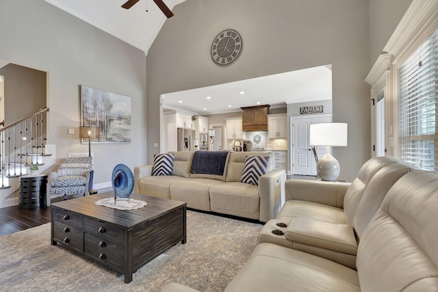 living room with ceiling fan, high vaulted ceiling, dark wood-type flooring, baseboards, and stairs