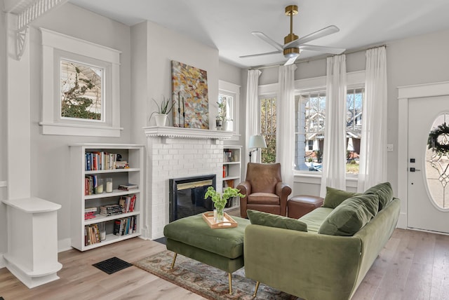 living area featuring a brick fireplace, ceiling fan, visible vents, and wood finished floors