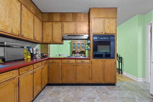 kitchen with brown cabinets, oven, a textured ceiling, stovetop, and under cabinet range hood