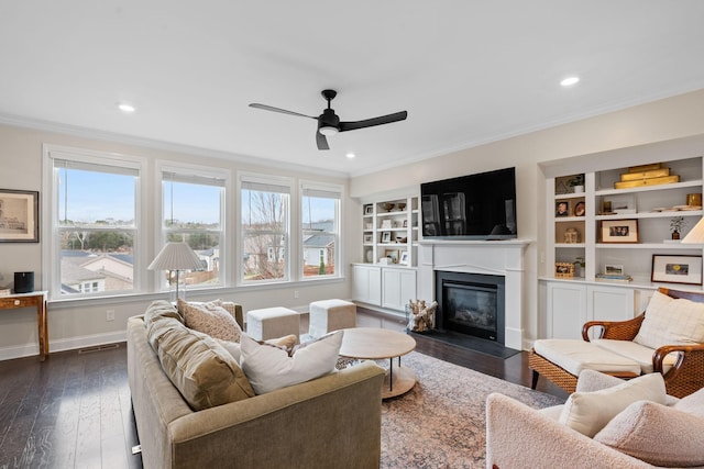 living area featuring baseboards, a glass covered fireplace, dark wood-style floors, ceiling fan, and ornamental molding