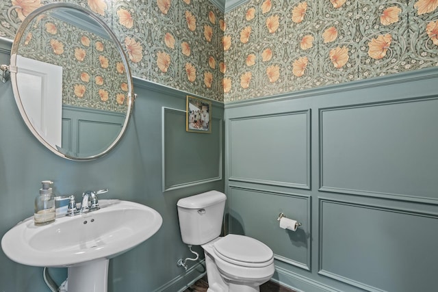 half bathroom with wallpapered walls, toilet, a wainscoted wall, a decorative wall, and a sink