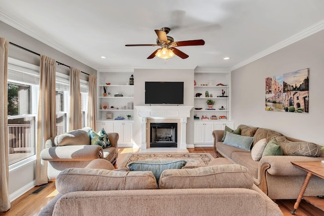 living room featuring built in features, crown molding, a fireplace with flush hearth, ceiling fan, and light wood-type flooring