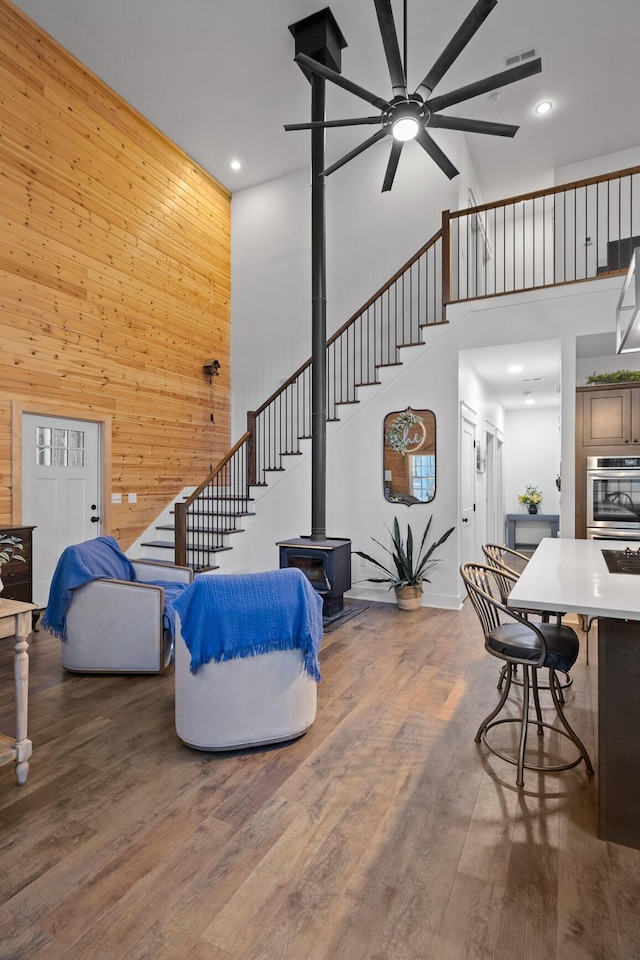 living room featuring a high ceiling, wood walls, wood finished floors, stairs, and a wood stove