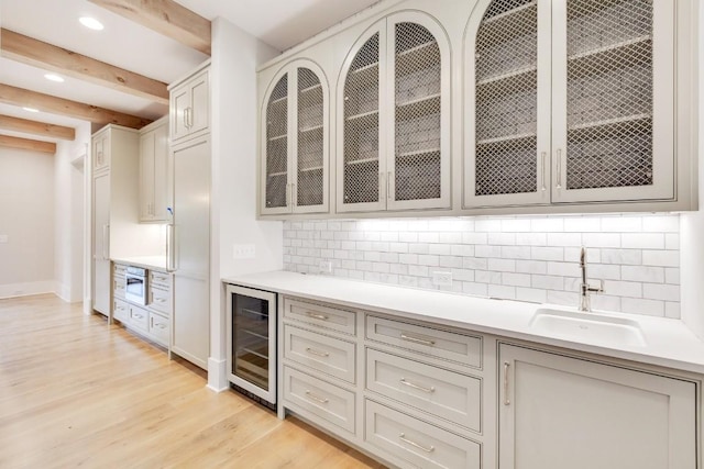 kitchen featuring glass insert cabinets, beverage cooler, beamed ceiling, light wood-style floors, and a sink