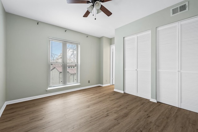 unfurnished bedroom featuring baseboards, visible vents, a ceiling fan, wood finished floors, and two closets