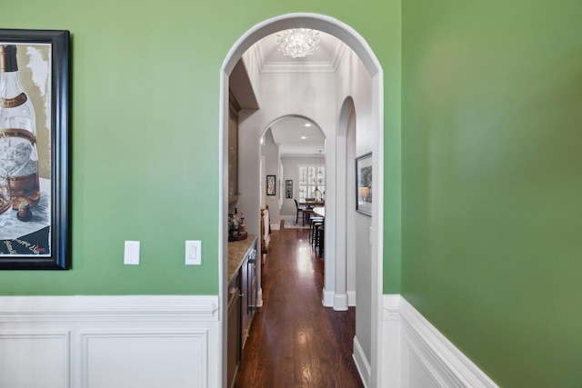 hallway with dark wood finished floors, arched walkways, an inviting chandelier, wainscoting, and crown molding