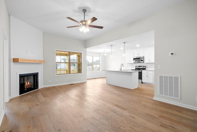 unfurnished living room featuring light wood-style flooring, ceiling fan with notable chandelier, a large fireplace, a sink, and visible vents