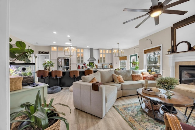 living room with recessed lighting, light wood finished floors, a tiled fireplace, and ceiling fan with notable chandelier