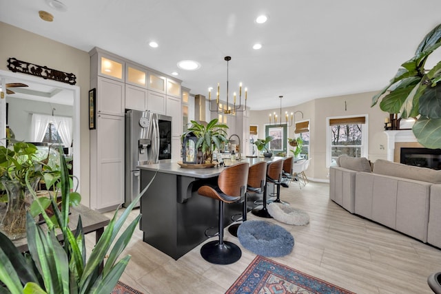 kitchen featuring a center island with sink, stainless steel refrigerator with ice dispenser, an inviting chandelier, glass insert cabinets, and a healthy amount of sunlight