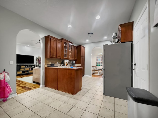 kitchen with arched walkways, glass insert cabinets, freestanding refrigerator, light tile patterned flooring, and a peninsula