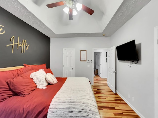 bedroom with ceiling fan, a textured ceiling, baseboards, light wood finished floors, and a tray ceiling