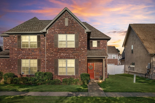 traditional-style house featuring a shingled roof, brick siding, fence, and a lawn