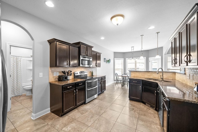 kitchen featuring arched walkways, stainless steel appliances, a sink, dark brown cabinets, and dark stone counters