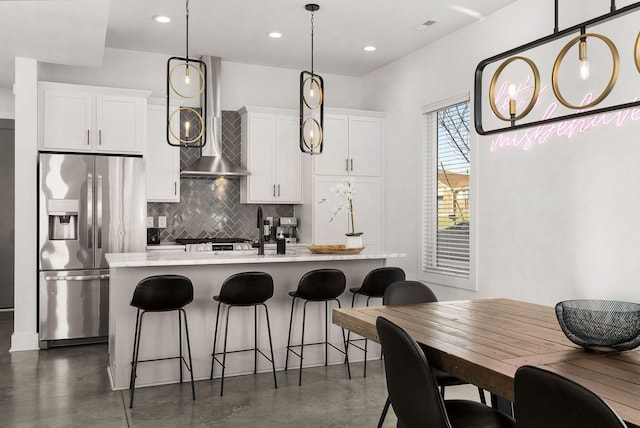 kitchen with white cabinets, light countertops, wall chimney range hood, stainless steel fridge with ice dispenser, and a center island with sink