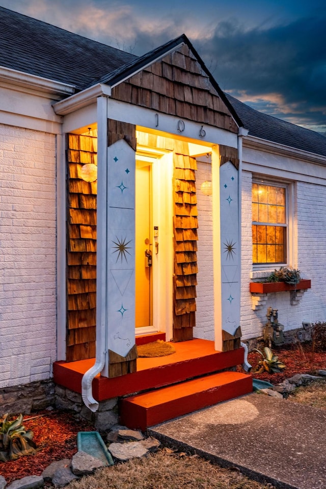 exterior entry at dusk featuring brick siding and a shingled roof