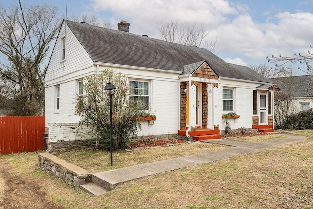 bungalow featuring brick siding, a chimney, roof with shingles, fence, and a front yard