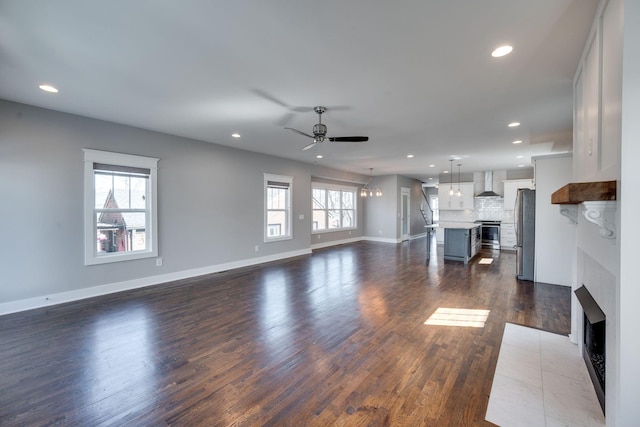 unfurnished living room featuring dark wood-style flooring, recessed lighting, a fireplace with flush hearth, baseboards, and ceiling fan with notable chandelier