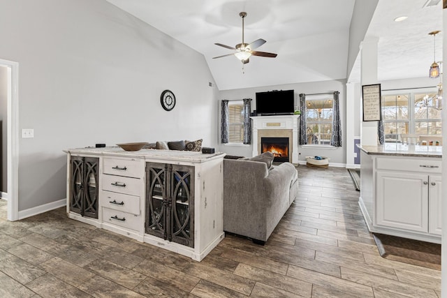 living room with vaulted ceiling, a warm lit fireplace, wood finish floors, and ceiling fan