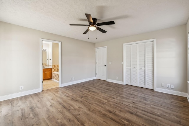 unfurnished bedroom featuring two closets, ensuite bathroom, a textured ceiling, wood finished floors, and baseboards