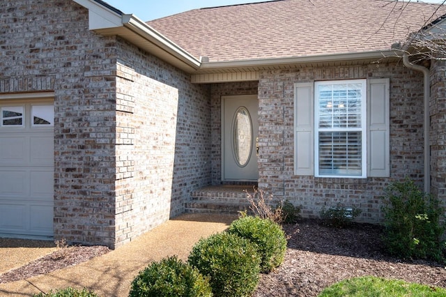 view of exterior entry featuring a garage, roof with shingles, and brick siding