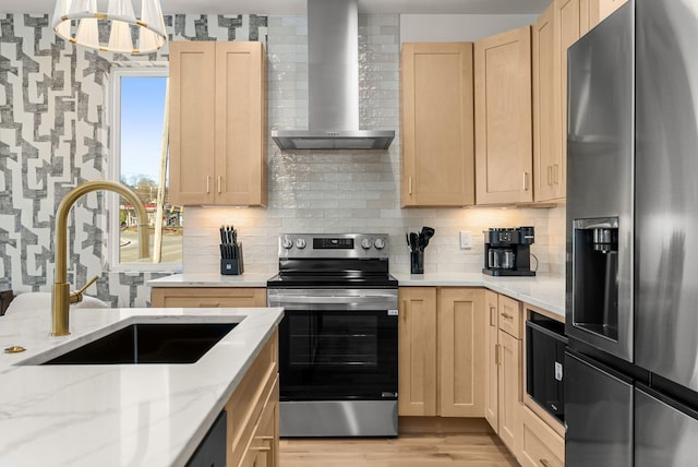 kitchen with light brown cabinetry, appliances with stainless steel finishes, a sink, wall chimney range hood, and wallpapered walls