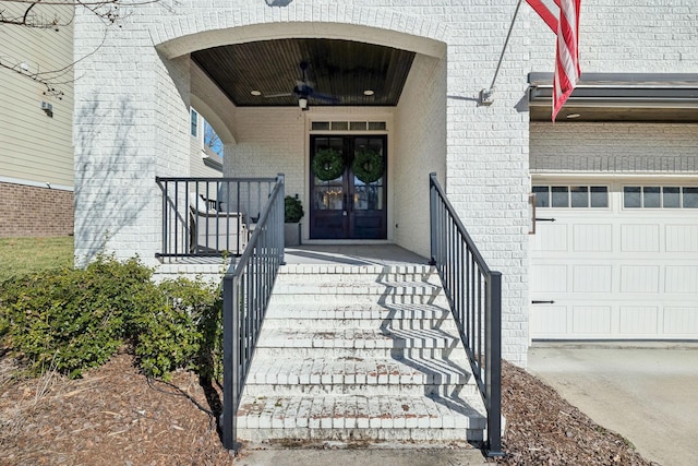 view of exterior entry with a garage, brick siding, ceiling fan, and french doors