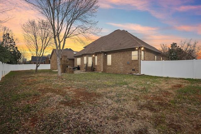 back of house at dusk featuring a fenced backyard, entry steps, crawl space, a lawn, and brick siding