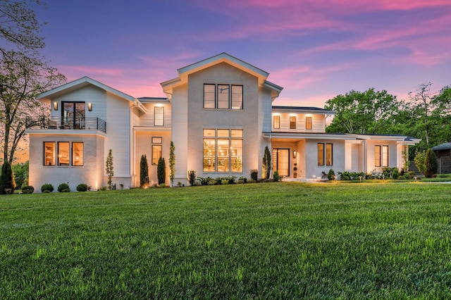 back of house at dusk featuring a lawn, a balcony, and stucco siding