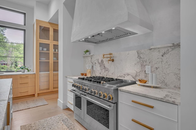 kitchen with light stone counters, tasteful backsplash, range with two ovens, light wood-type flooring, and wall chimney exhaust hood