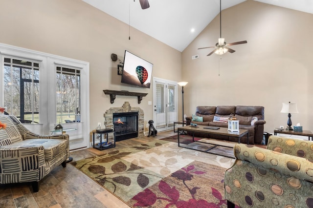 living area with ceiling fan, high vaulted ceiling, a stone fireplace, wood finished floors, and visible vents