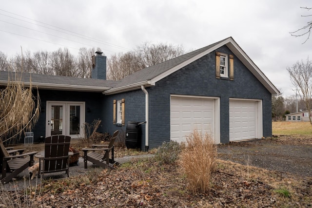 rear view of property featuring a garage, brick siding, driveway, french doors, and a chimney