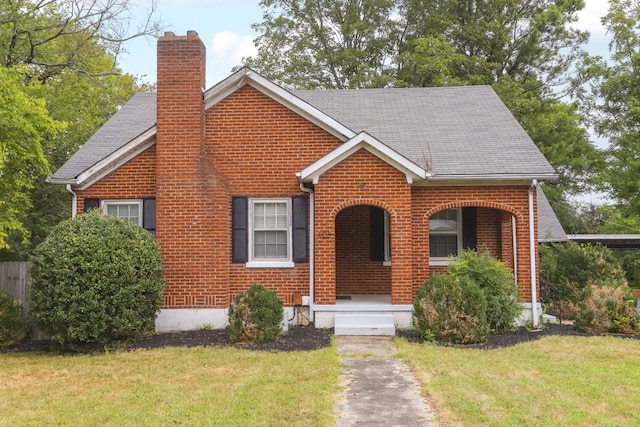 view of front facade featuring brick siding, a chimney, a front yard, and a shingled roof