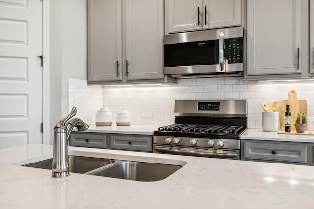 kitchen featuring gray cabinetry, light stone countertops, decorative backsplash, appliances with stainless steel finishes, and a sink