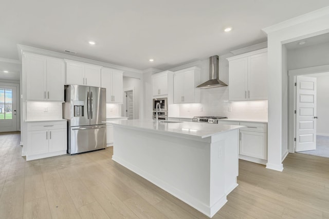 kitchen with a kitchen island with sink, light wood-style flooring, stainless steel appliances, white cabinetry, and wall chimney range hood