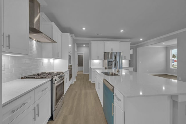 kitchen with a sink, appliances with stainless steel finishes, wall chimney exhaust hood, light wood finished floors, and an island with sink