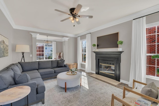 living room with ceiling fan with notable chandelier, wood finished floors, a fireplace with flush hearth, baseboards, and ornamental molding