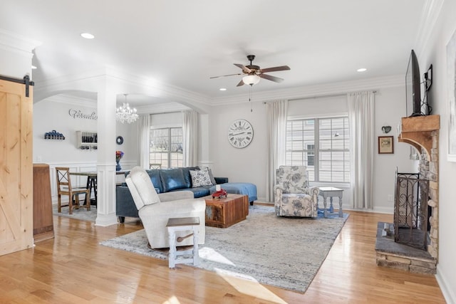 living room with crown molding, light wood finished floors, a fireplace with raised hearth, a barn door, and ceiling fan with notable chandelier