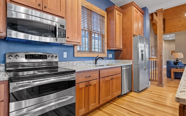 kitchen featuring light wood-style flooring, appliances with stainless steel finishes, brown cabinetry, a sink, and light stone countertops