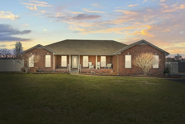 back of property at dusk with a shingled roof, brick siding, a yard, and fence