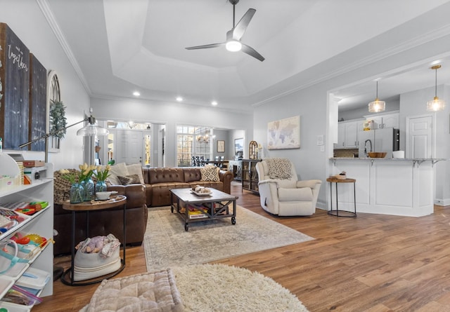 living room with ornamental molding, wood finished floors, ceiling fan with notable chandelier, and a raised ceiling