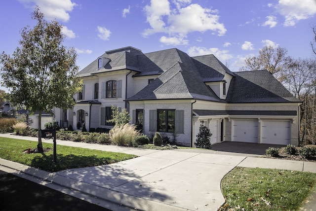 french country inspired facade with roof with shingles, brick siding, an attached garage, a front yard, and driveway