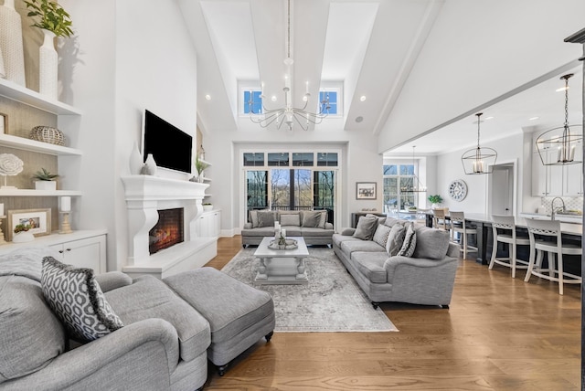 living room featuring high vaulted ceiling, wood finished floors, a lit fireplace, and an inviting chandelier