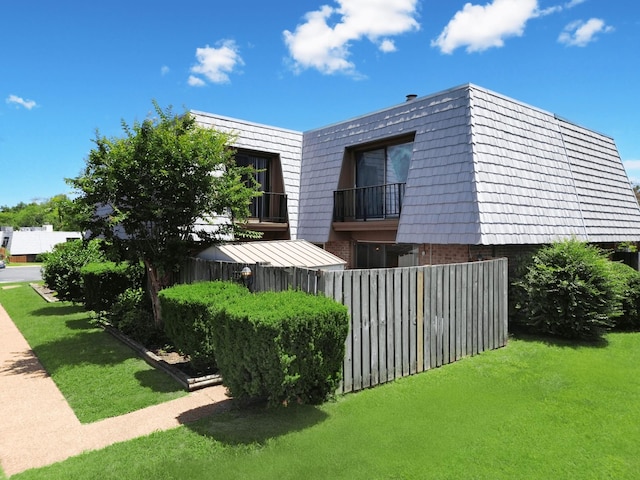 view of side of home featuring mansard roof, a balcony, brick siding, fence, and a yard