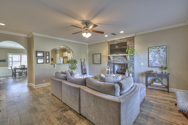 living area with ornamental molding, a ceiling fan, wood finished floors, arched walkways, and a stone fireplace