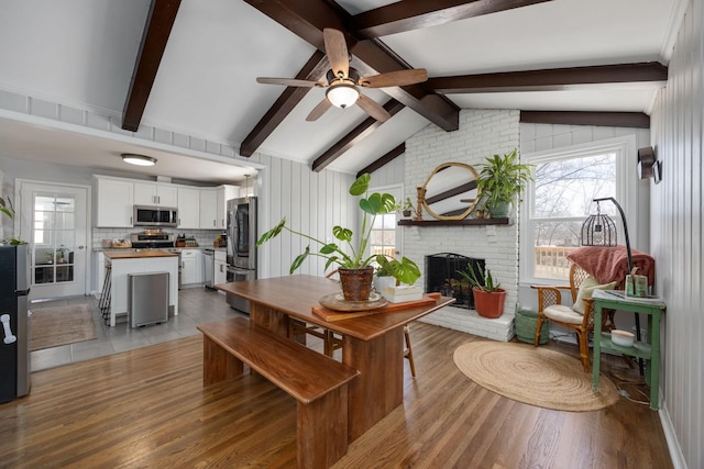 dining room with a brick fireplace, ceiling fan, vaulted ceiling with beams, and light wood finished floors