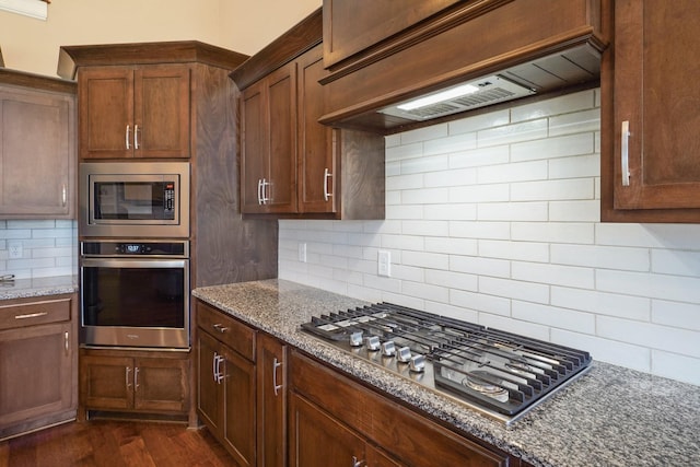 kitchen featuring custom exhaust hood, appliances with stainless steel finishes, dark stone counters, and dark wood finished floors